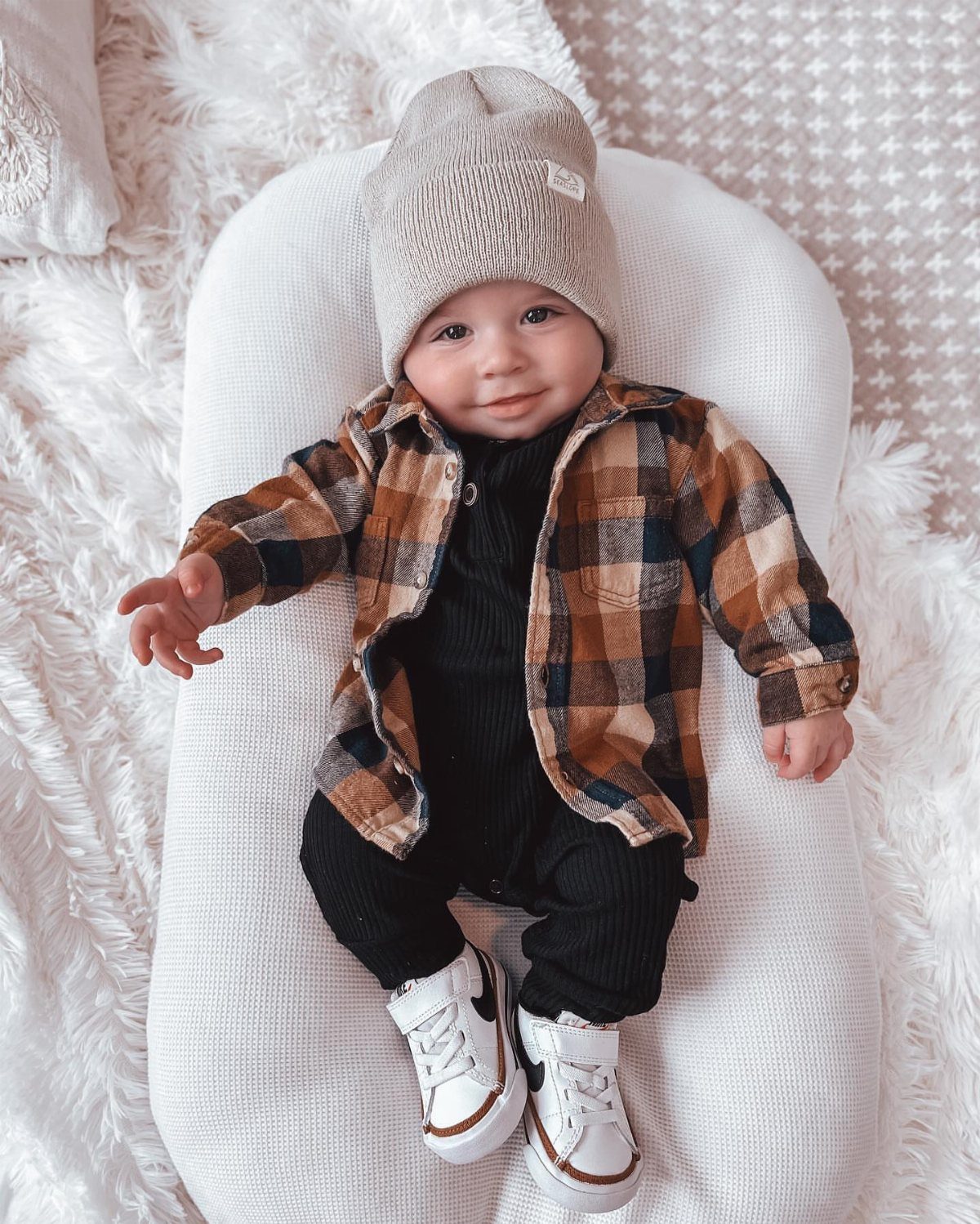 Baby boy in a plaid shirt and knit beanie lying on white fur.