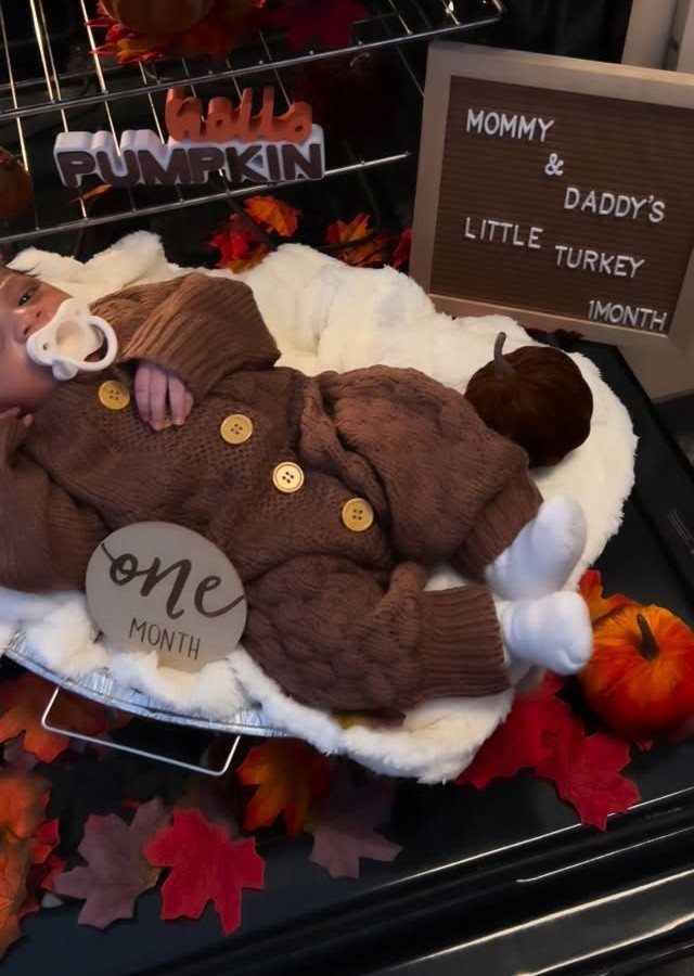 Newborn baby in brown knit romper surrounded by autumn leaves and pumpkins.