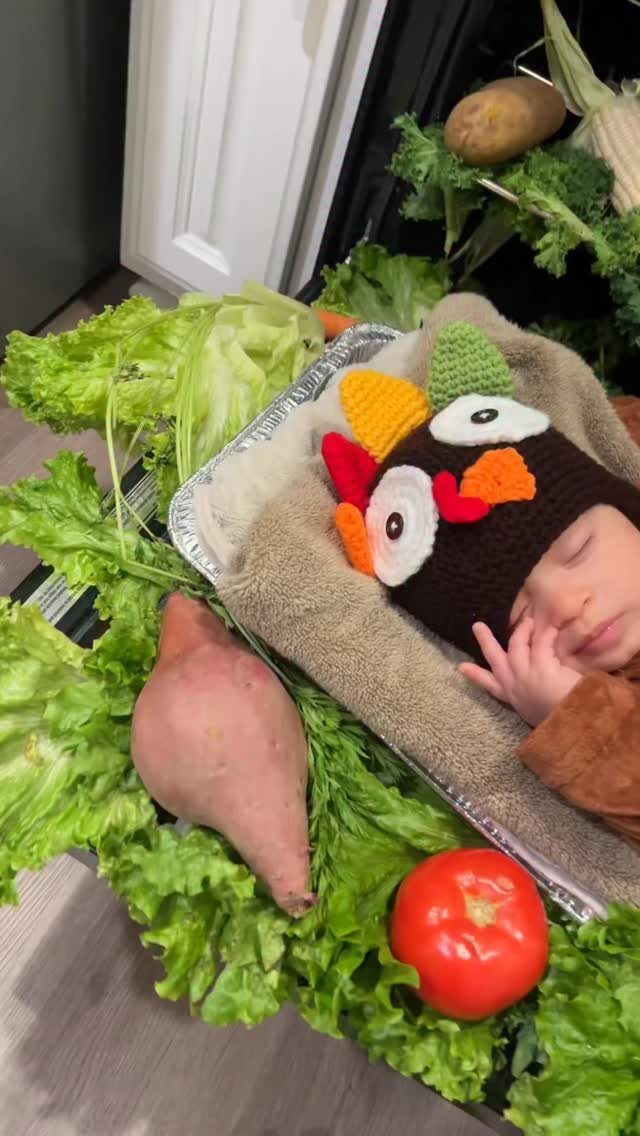 Newborn baby wearing a crocheted turkey hat lying in a vegetable display