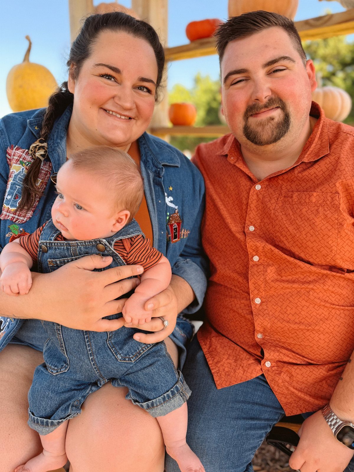 Newborn baby in denim overalls and orange shirt with parents and pumpkins.