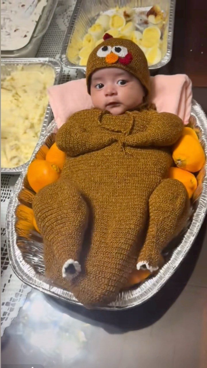 Newborn baby in a brown crocheted turkey costume lying in a foil tray.