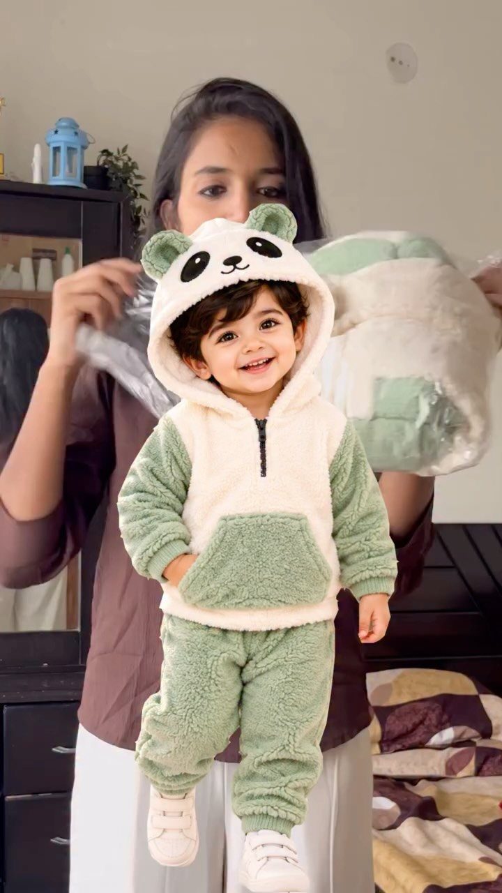 Infant wearing a fuzzy light green and white panda hooded tracksuit and white shoes.