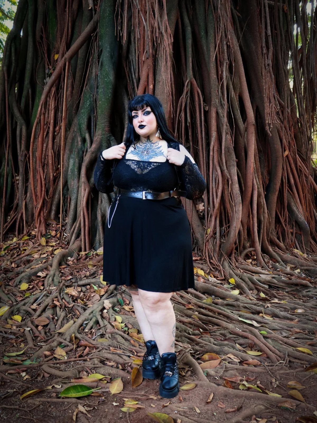 Plus-size woman in a black gothic lace top and skirt standing by tree roots.