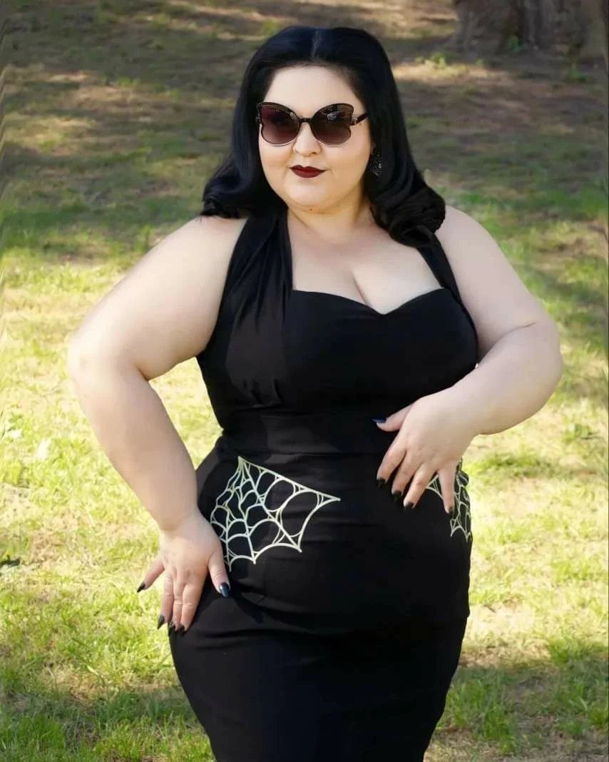 Plus-size woman in a black halter dress with spiderweb details posing on grass.