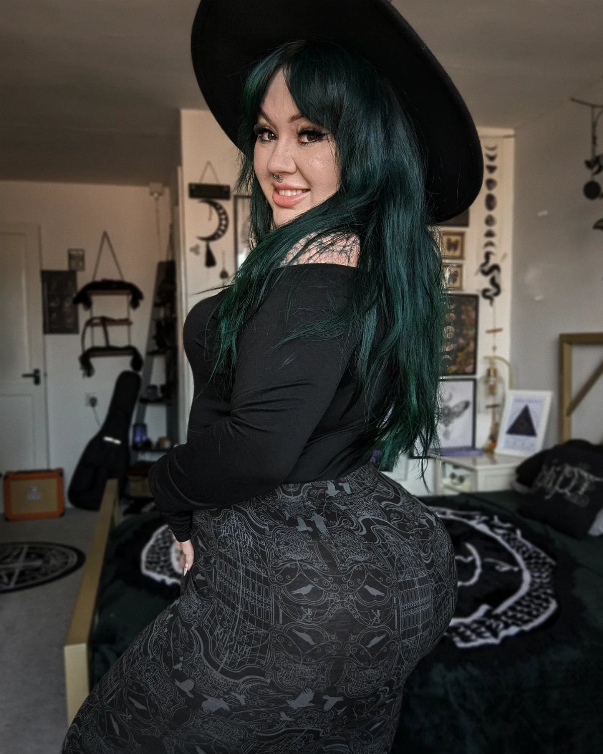 Woman with green hair in gothic attire posing in a decorated bedroom.