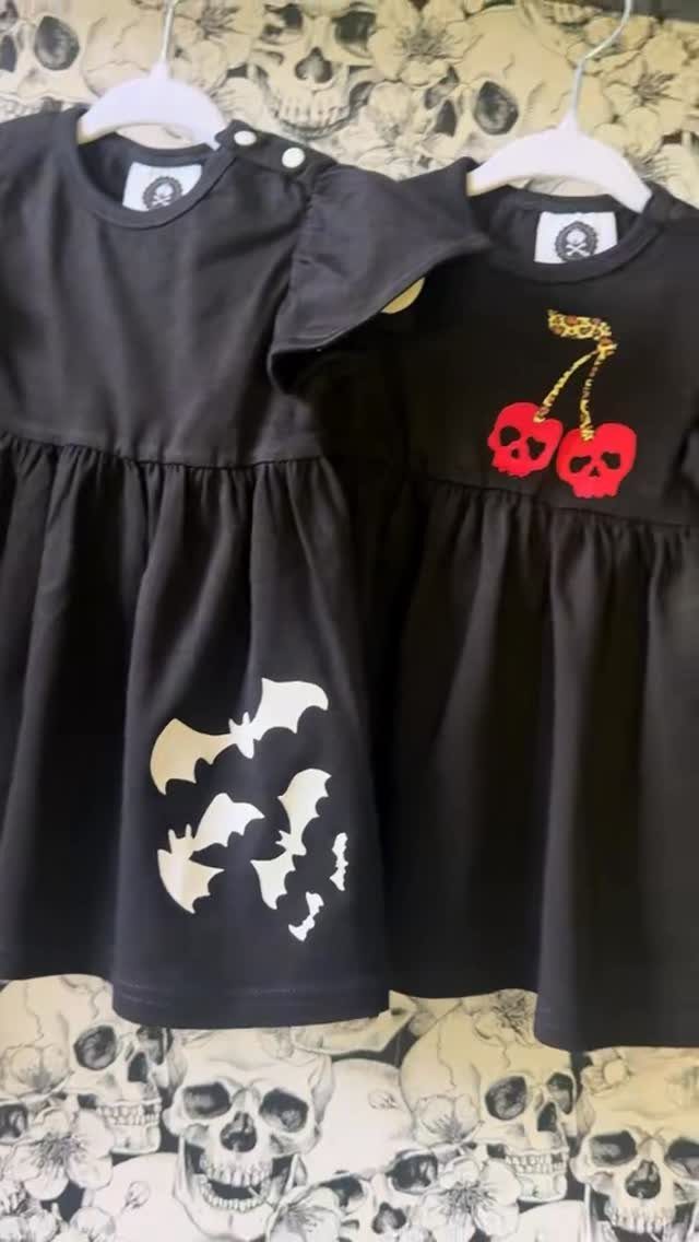 10 Rockabilly Infant Clothing Ideas for Little Rock Stars 9 Two black infant dresses with bat and skull cherry graphics on skull wallpaper.