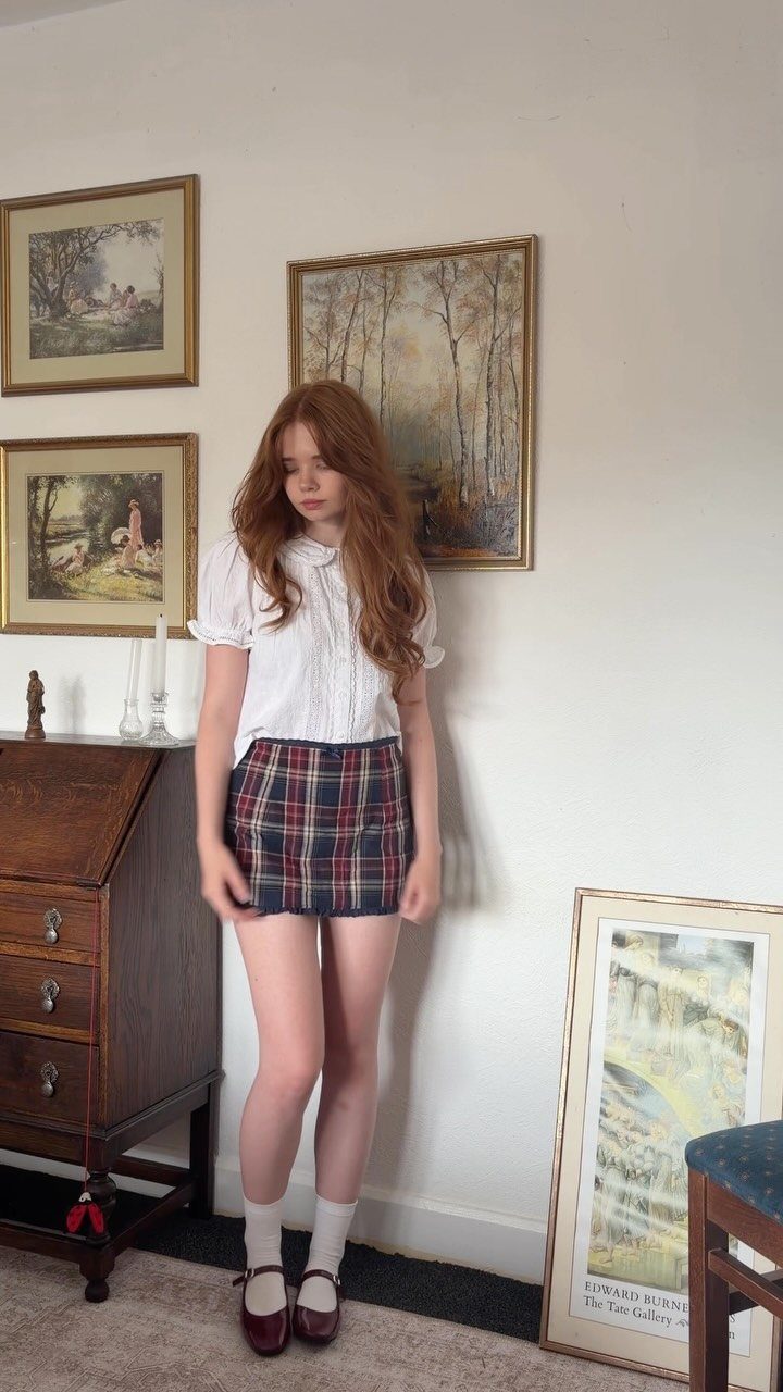 Woman with red hair wearing a white blouse and plaid mini skirt indoors.