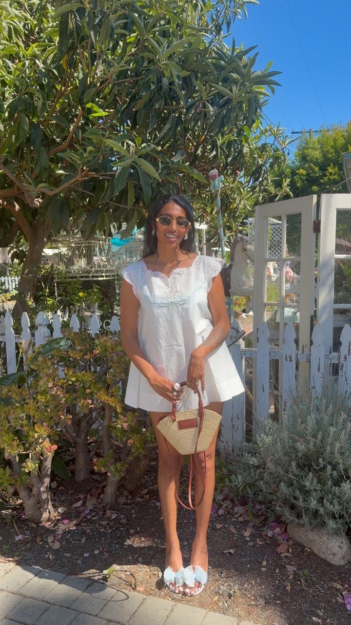 Woman in a short white lace dress holding a straw bag outdoors.