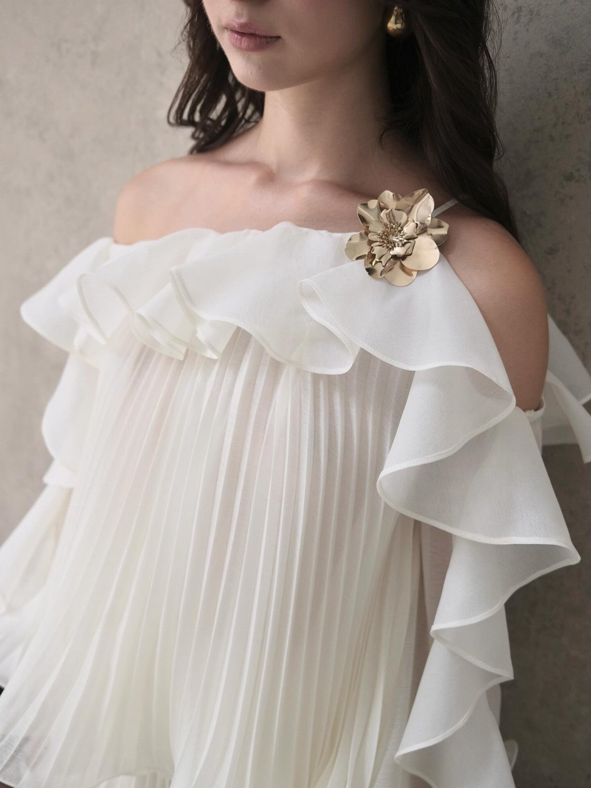 Woman wearing a white pleated off-the-shoulder dress with ruffles and gold flower brooch.