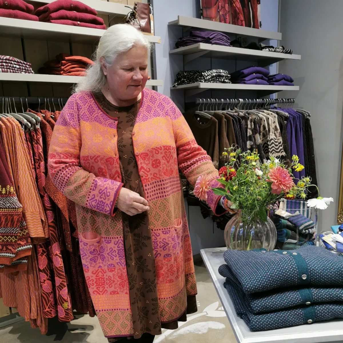 Swedish Women's Clothing Ideas: 8 Outfits to Wear Now 1 Woman in a patterned orange cardigan standing in a clothing store with flowers.