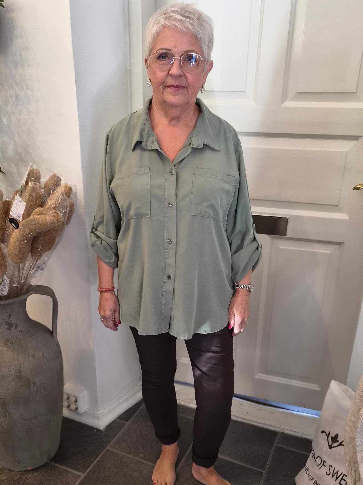 Swedish Women's Clothing Ideas: 8 Outfits to Wear Now 7 Older woman wearing a sage green shirt and black leather leggings in a hallway.