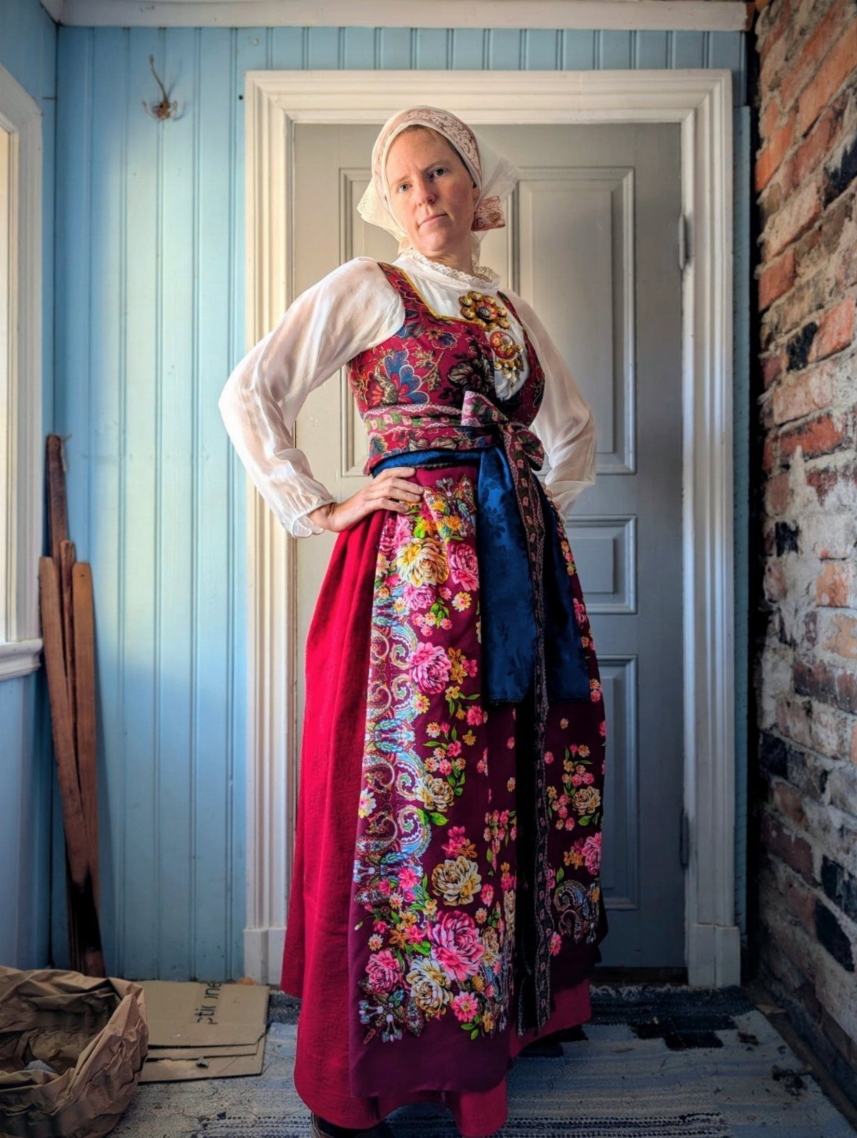 Woman in traditional Danish costume with floral apron and white bonnet standing indoors.
