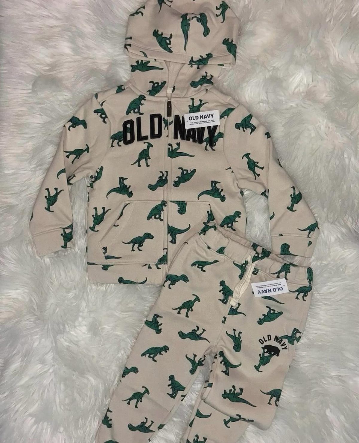 Beige baby tracksuit with green dinosaur patterns on a white faux-fur background.