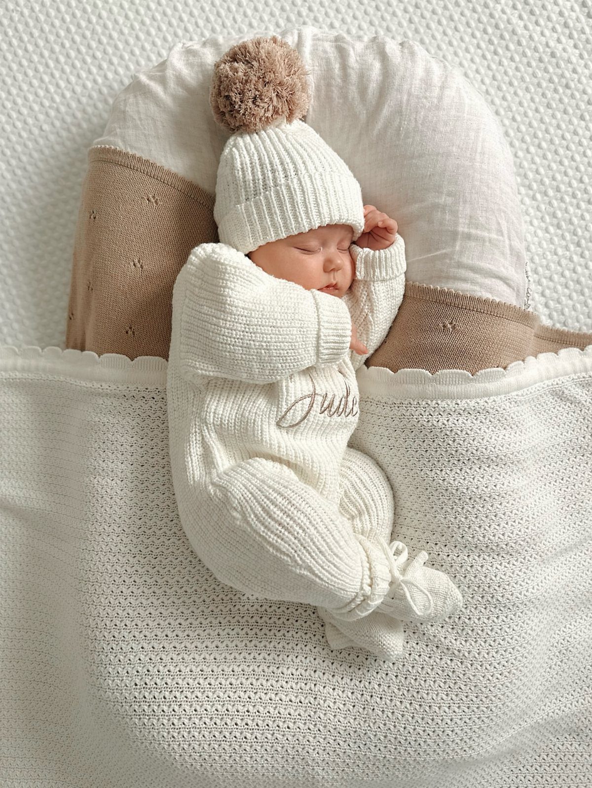 Sleeping newborn baby in a white knitted outfit and pom-pom beanie on a blanket.