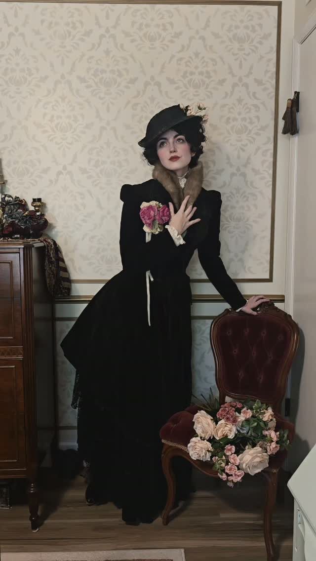 Woman in black Victorian era clothing and hat standing in a vintage room.