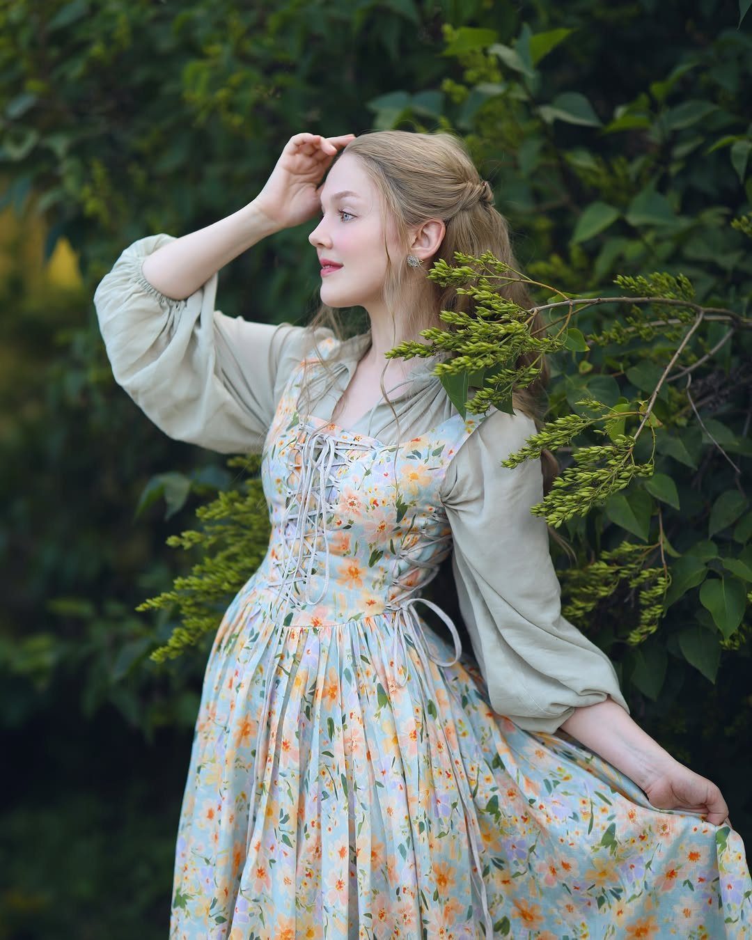 Woman in a floral Victorian style dress and green blouse standing by greenery.