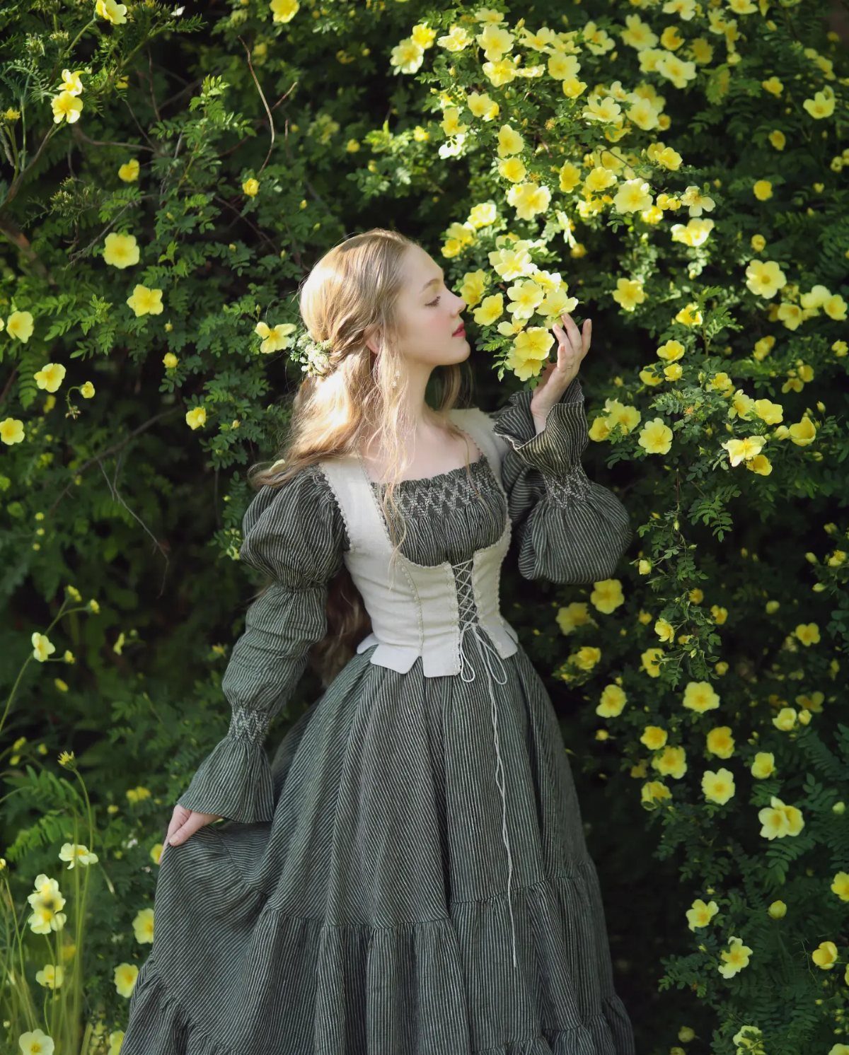 Woman in a Victorian-style olive green dress smelling yellow flowers in a garden