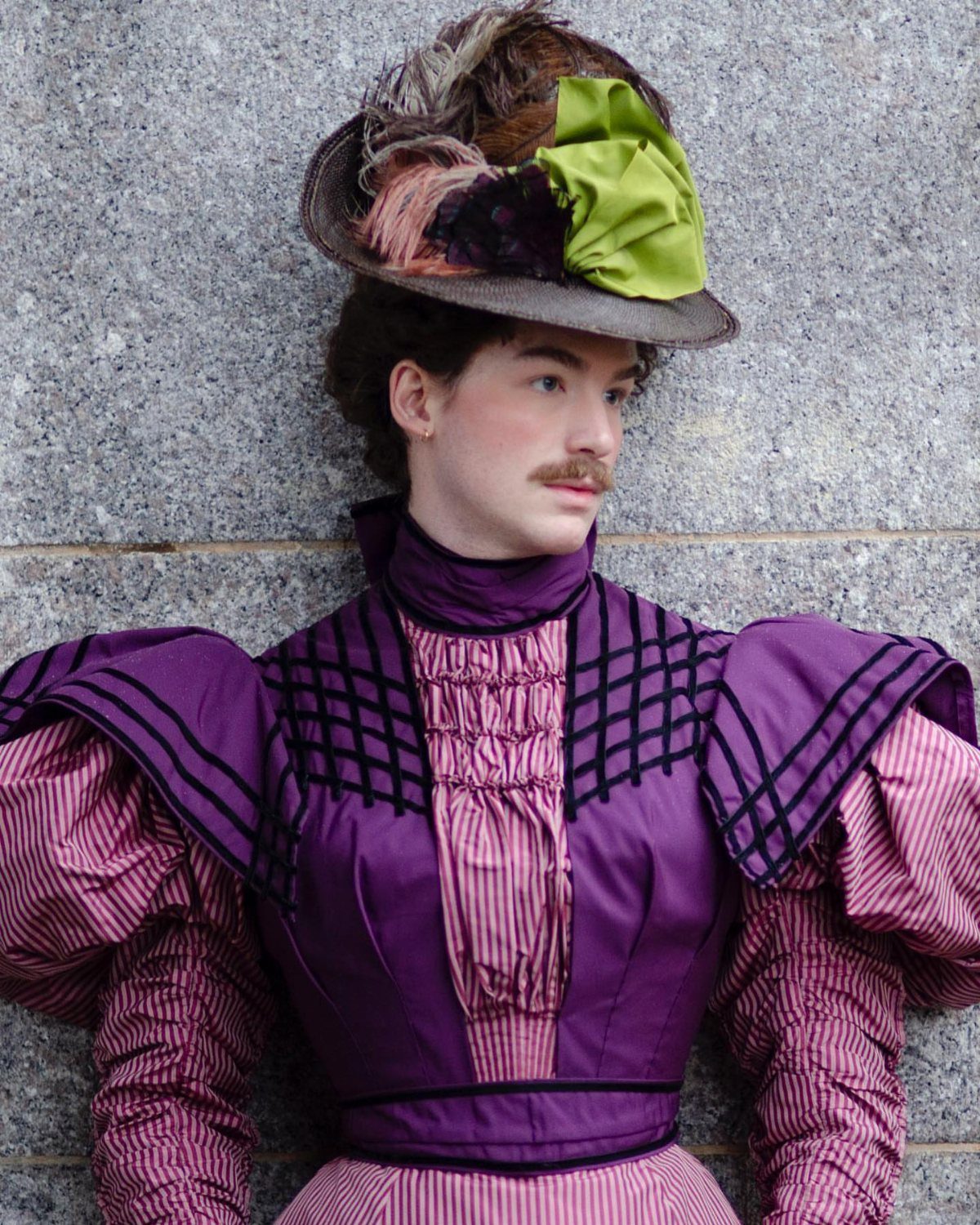 Person in purple Victorian era clothing and a decorative hat against stone wall