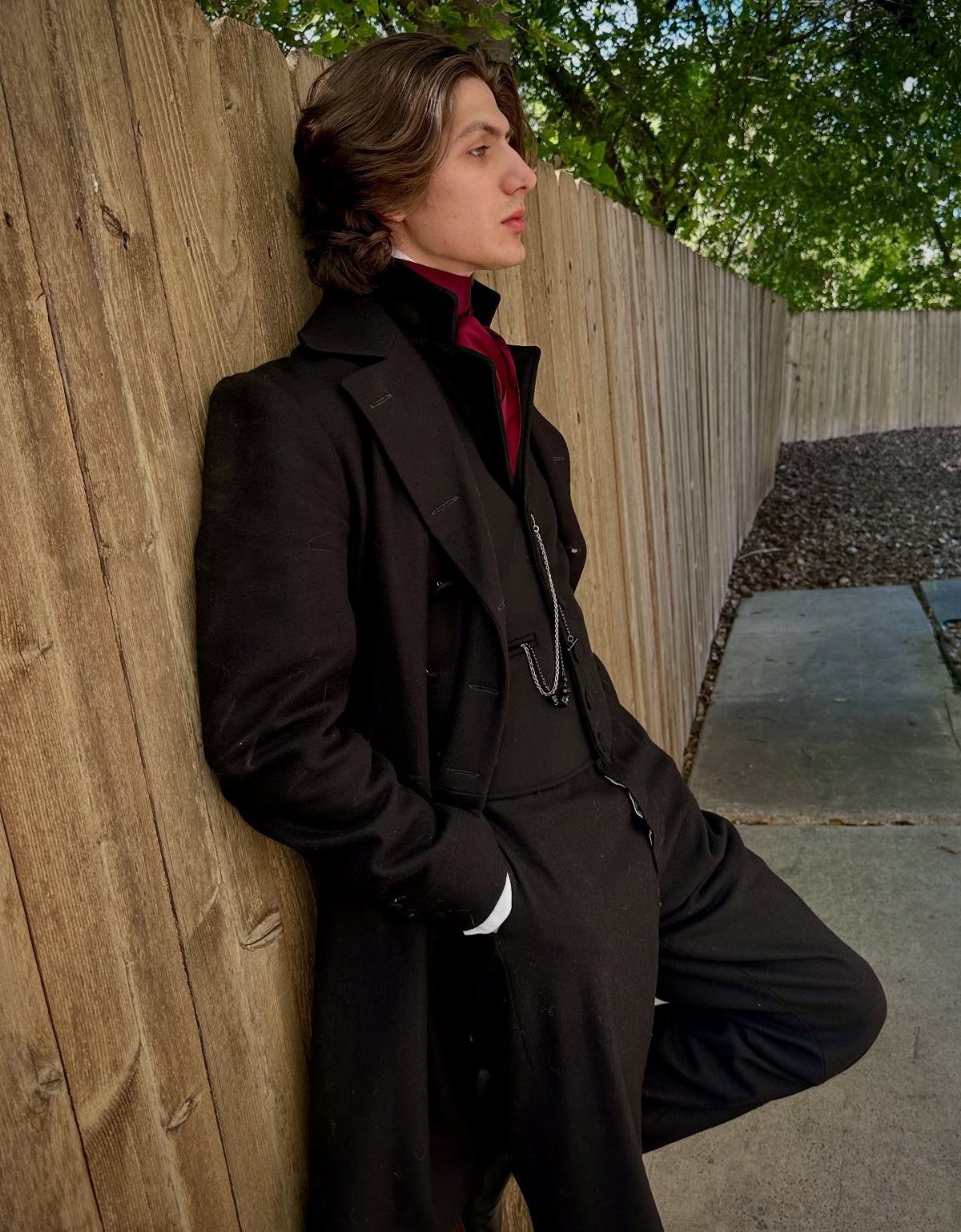 Man in black Victorian frock coat and red cravat leaning on wooden fence
